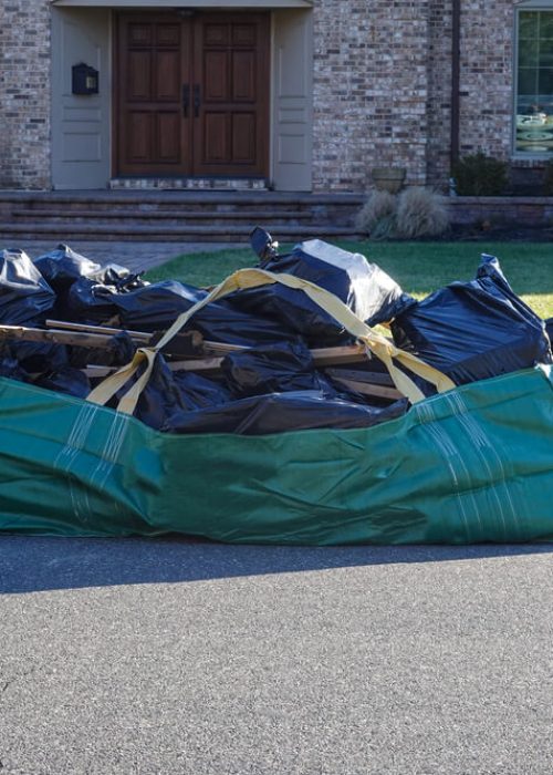Green large trash bag filled with black plastic garbage bags on an asphalt street near the curb in front of a large house with a big green lawn.